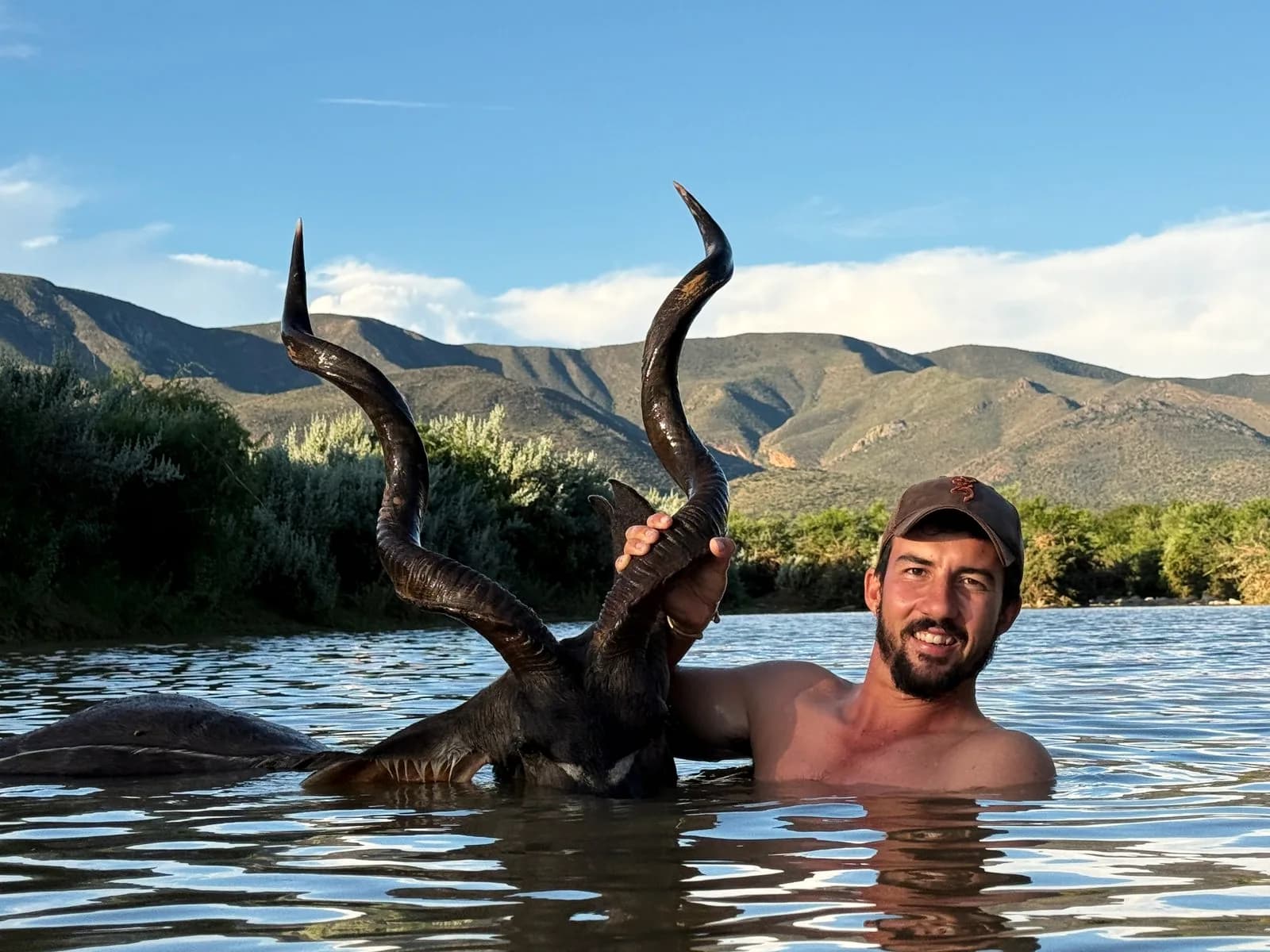 A hunter recovering a kudu bull from a river below the mountains